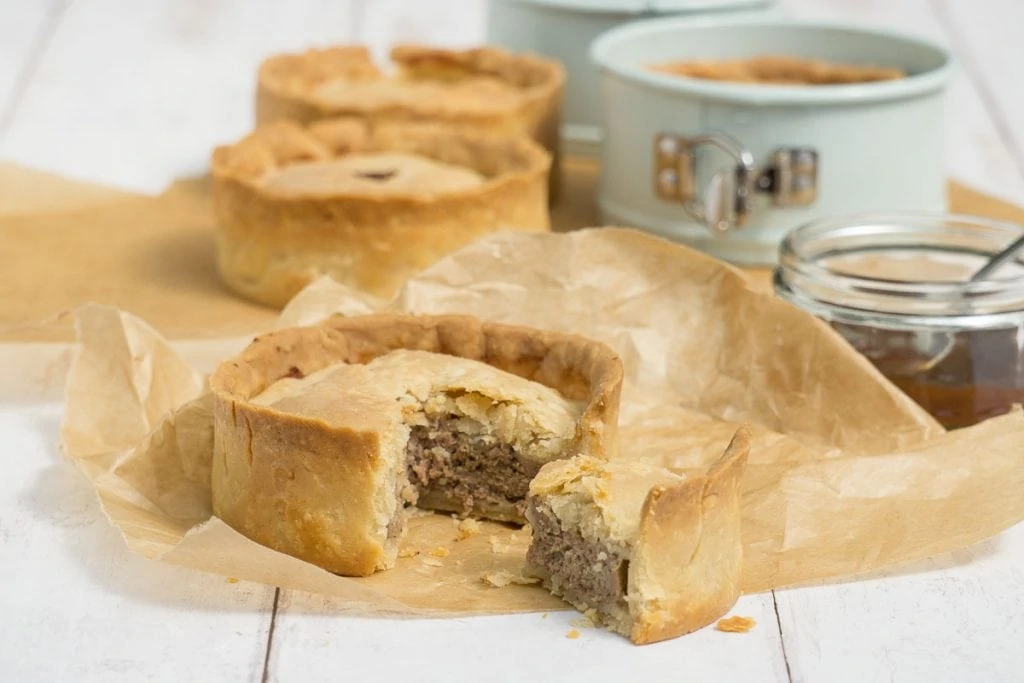 Scotch Pie Recipe - Scotch pie with slice cut out on brown paper and other pies and tin in the background 

