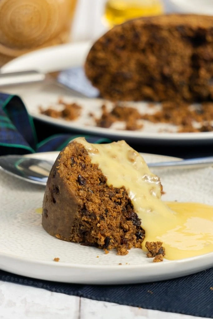 Scottish Clootie Dumpling Recipe on a plate with custard and whole pudding in the background