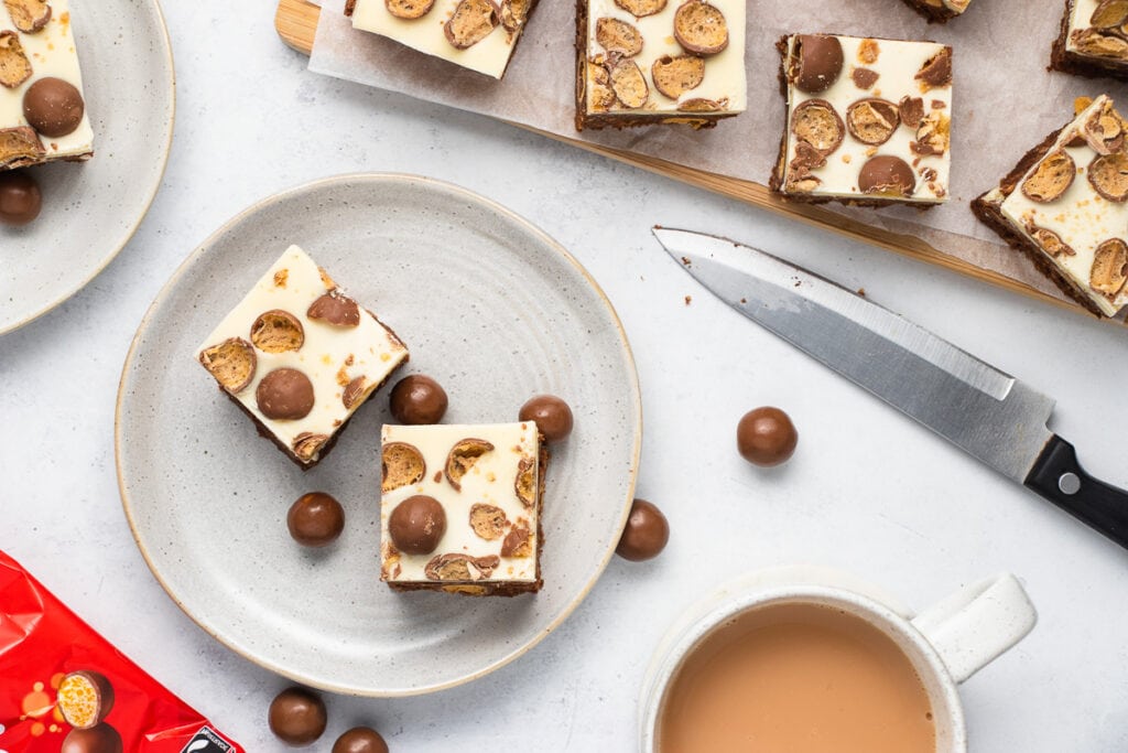 Malteser Traybake Recipe - Slices on a plate, a cup of tea, and more slices on a tray with a knife and Malteser packet nearby