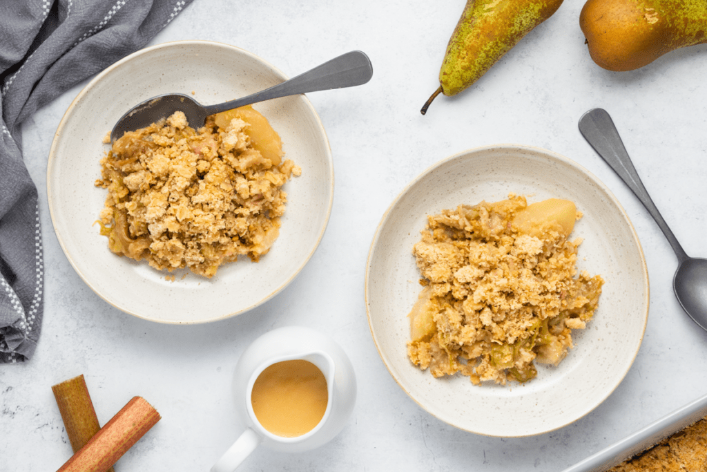 Rhubarb and Pear Crumble Recipe - Two bowls of crumble next to a tea towel, spoons, pear and rhubarb