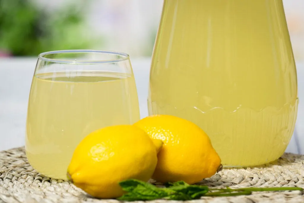 Homemade Lemon Cordial Recipe - two lemons with glass and jug of cordial behind